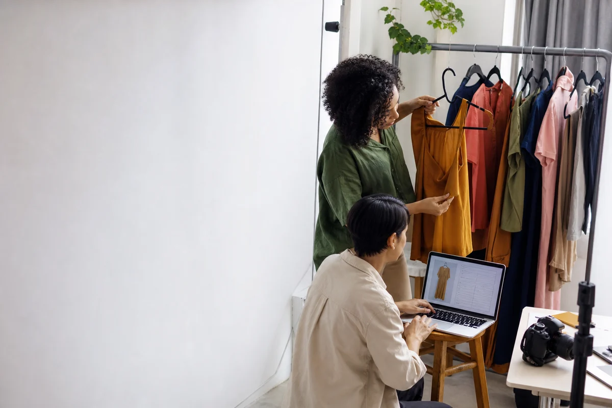 Fotógrafo de produto e cadastrador trabalhando juntos num canto de estúdio, roupas na arara e um notebook com uma ficha sendo preenchida